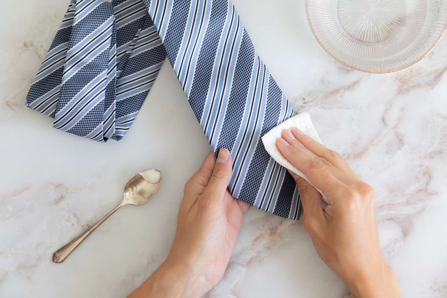 A damp cloth is being pressed against the necktie to eliminate any solid particles, situated beside a spoon and a glass bowl filled with water.