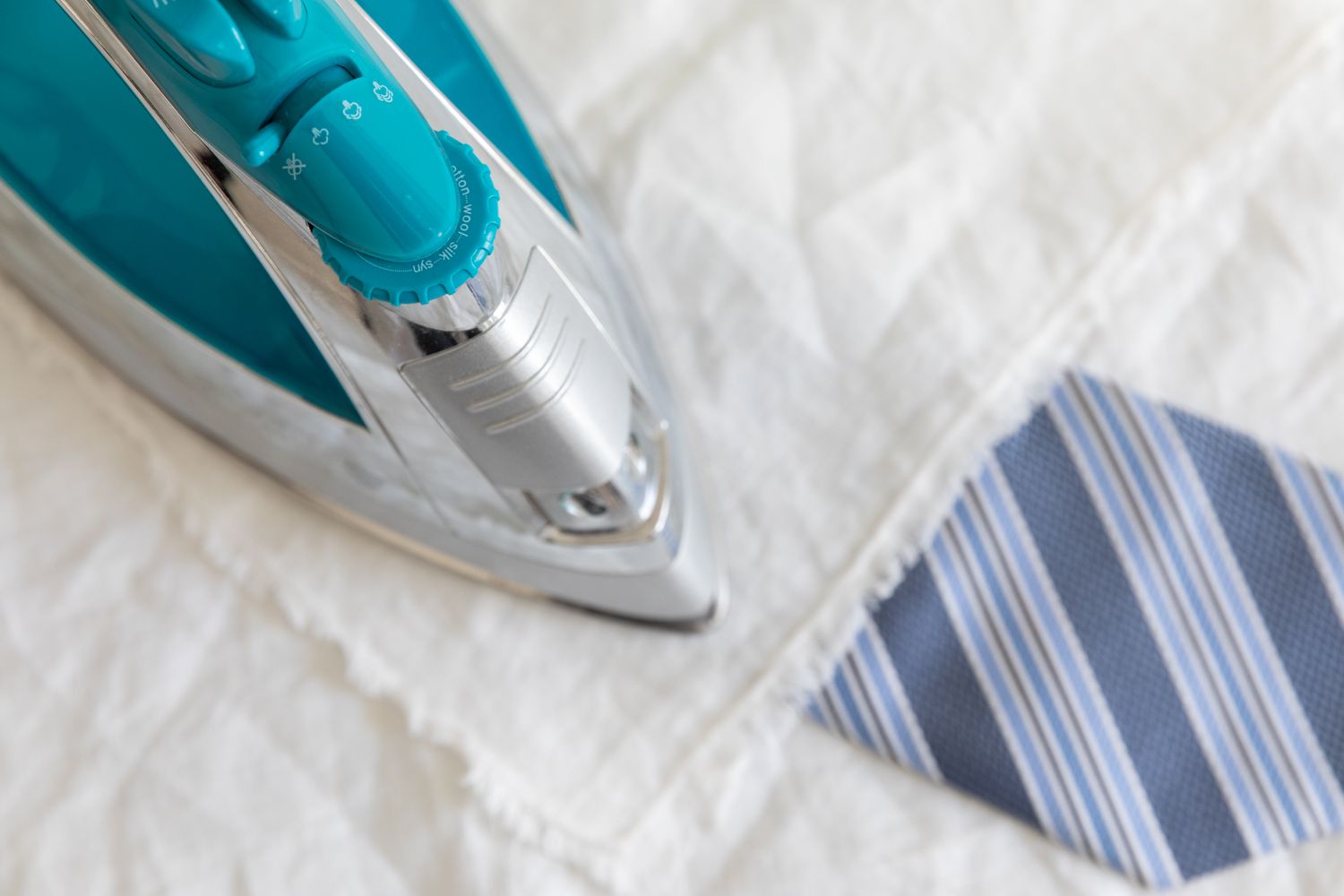 A blue necktie is being pressed using a teal steam iron, with a pressing cloth placed in between.