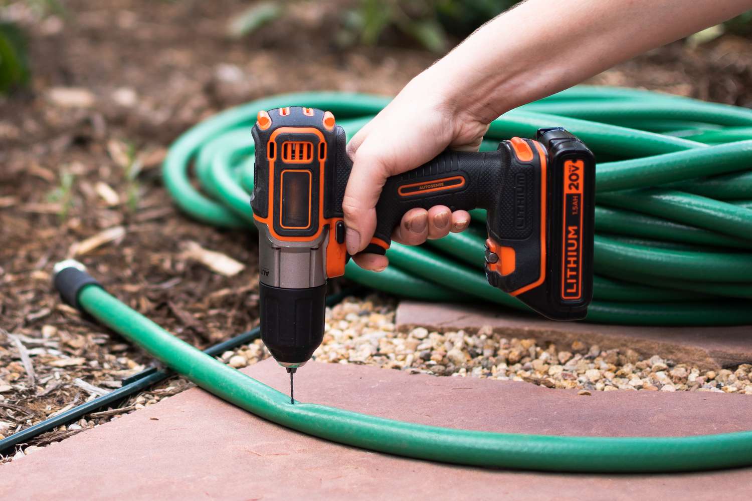 An orange power drill making openings in the side of a green garden hose.